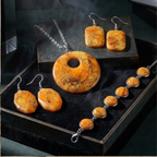 "A Diddit Amber jewellery set displayed in a black presentation box, featuring a round pendant necklace, a beaded bracelet, and matching earrings crafted from polished amber-coloured stone with metallic gold flecks, suspended from silver-tone chains."