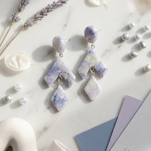 "A pair of Diddit Amethyst drop earrings featuring irregular lavender, purple, and white marbled stone pieces on silver hooks, styled on a white marble surface with decorative pearls, dried lavender, and small white accents."