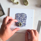 "A pair of hands using a small clay cutter to cut a decorative Diddit Amethyst patterned clay piece from a white board, with additional clay cutters visible on a light marble surface."