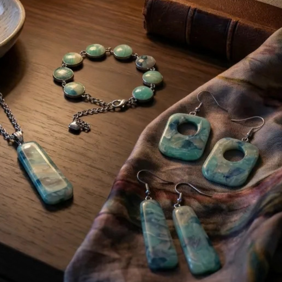"A Diddit Aquamarine jewellery set featuring a silver pendant necklace, a beaded bracelet, and matching earrings with turquoise-blue marbled stones, displayed on a wooden surface with a patterned cloth."