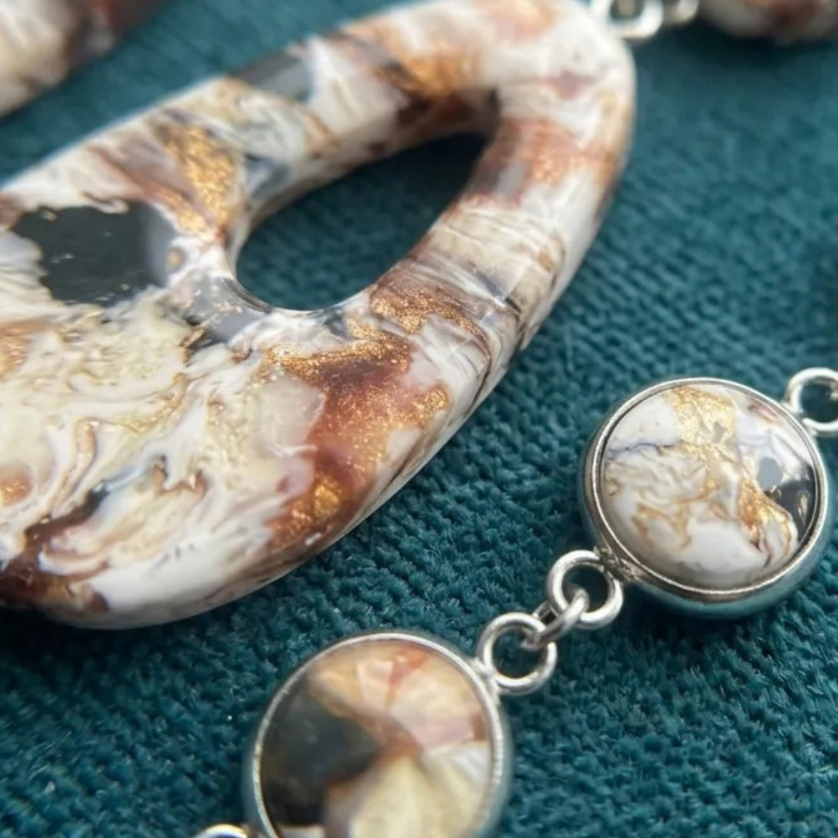 "Close-up of a Diddit Desert Onyx teardrop pendant necklace with matching round drop charms, featuring a marbled white, brown, and black pattern with metallic gold flecks, set in silver-tone metal on a textured teal background."