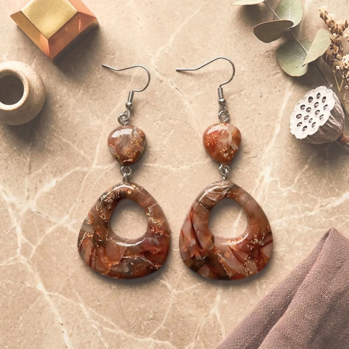 "A pair of Diddit Desert Onyx teardrop drop earrings featuring large brown and orange marbled stone pendants, a smaller matching stone detail, and silver fishhook wires, styled on a beige marble surface with dried botanicals and fabric."
