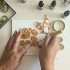 "A pair of hands using a blade tool to cut and shape Diddit Amber orange and brown marbled polymer clay pieces in various circle, oval, and square shapes on a white tile, surrounded by olive-green geometric clay cutters, clay offcuts, and Diddit alcohol ink bottles on a white marble surface."