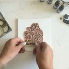"A pair of hands pressing an olive-green clay cutter into a Diddit Tortoiseshell marbled orange, brown, and black patterned polymer clay sheet on a white tile, surrounded by a box of olive-green clay cutters, Diddit alcohol ink bottles, and clay scraps on a white marble surface."