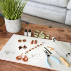 "A Diddit Tortoiseshell jewellery making experience kit laid out on a white tile on a wooden table, featuring orange and brown marbled clay earrings in multiple styles, a beaded bracelet, blue-handled pliers, an acrylic rolling pin, gold leaf pieces, silver jewellery findings, olive-green clay cutters, and Diddit alcohol ink bottles.