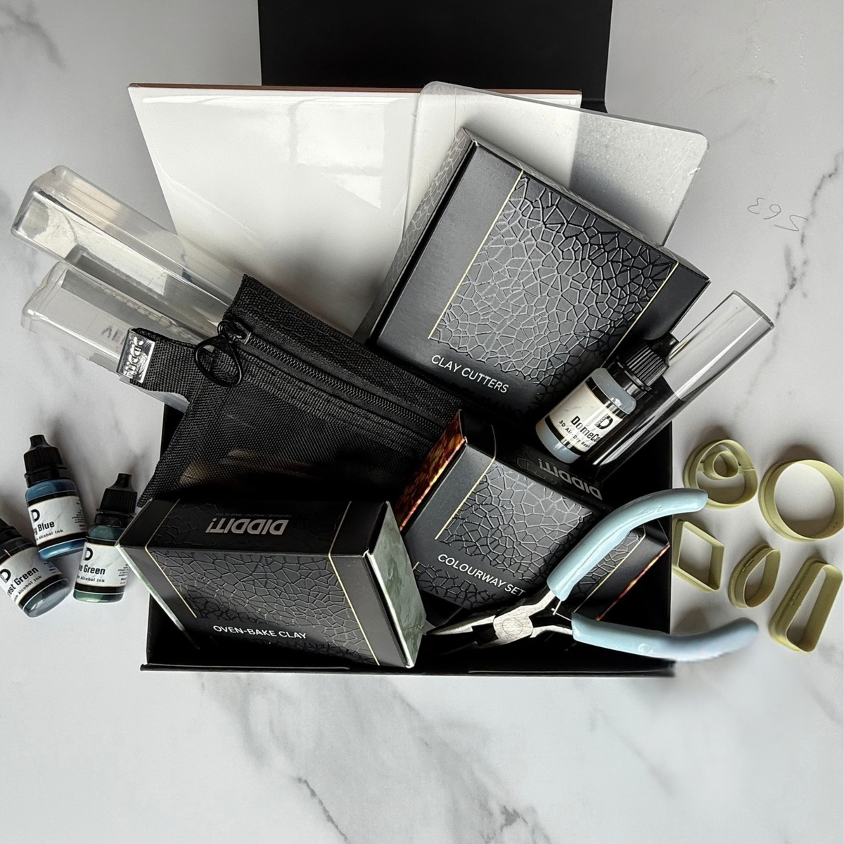 "An overhead flat-lay of a Diddit Amber jewellery making experience kit displayed in an open black box on a white marble surface, featuring a Diddit oven-bake clay box, clay cutters box, colourway set box, black zip bag with tools, alcohol ink bottles, blue-handled pliers, olive-green geometric clay cutters, an acrylic rolling pin, and a backing tile."