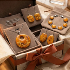 "A Diddit Amber jewellery gift set displayed in an open presentation box with a burnt orange satin ribbon, featuring a round pendant necklace, two rectangular drop earrings with gold flecks, two oval drop earrings, and a silver chain bracelet with round amber cabochons, presented on taupe velvet inserts."