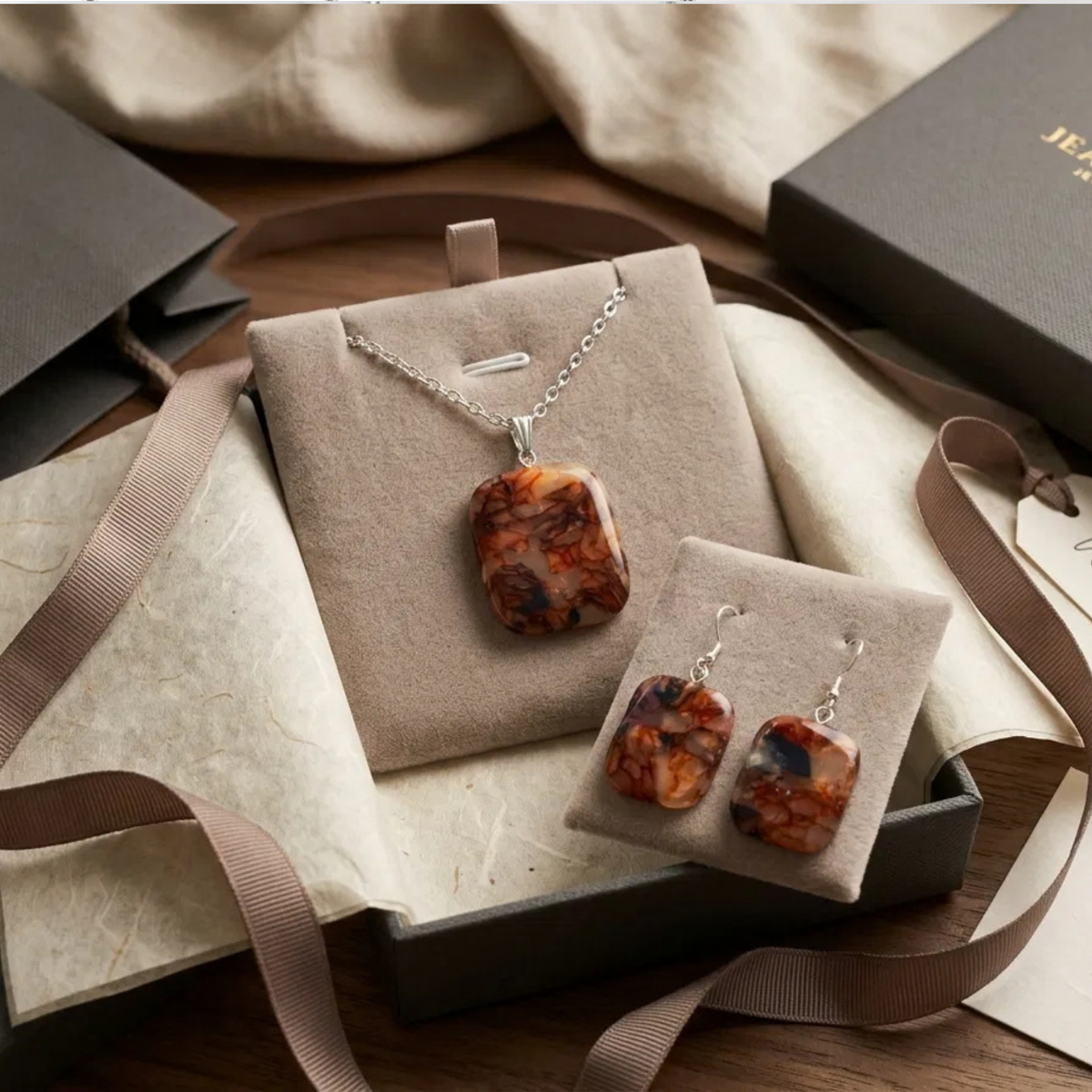 "A Diddit Tortoiseshell jewellery set featuring a pendant necklace and matching drop earrings with marbled amber-brown and black stones on silver chains, displayed on beige velvet jewellery stands inside an open gift box with ribbon and packaging."

