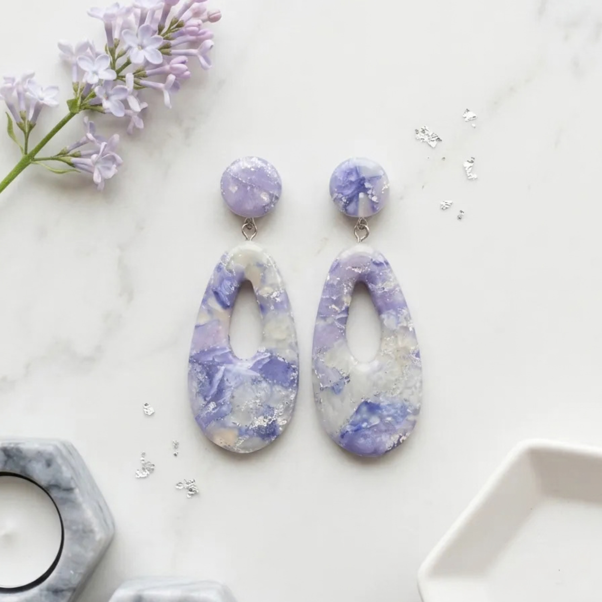 "A pair of Diddit Amethyst teardrop drop earrings with a marbled lavender, purple, and white pattern accented with silver foil flakes, displayed on a white marble surface with decorative flowers and small silver accents."