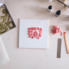 A top-down view of a white ceramic tile with polymer clay pieces arranged on it, surrounded by crafting tools and bottles on a light marble-like surface. Diddit's Rose Quartz Jewellery Making Craft Kit.