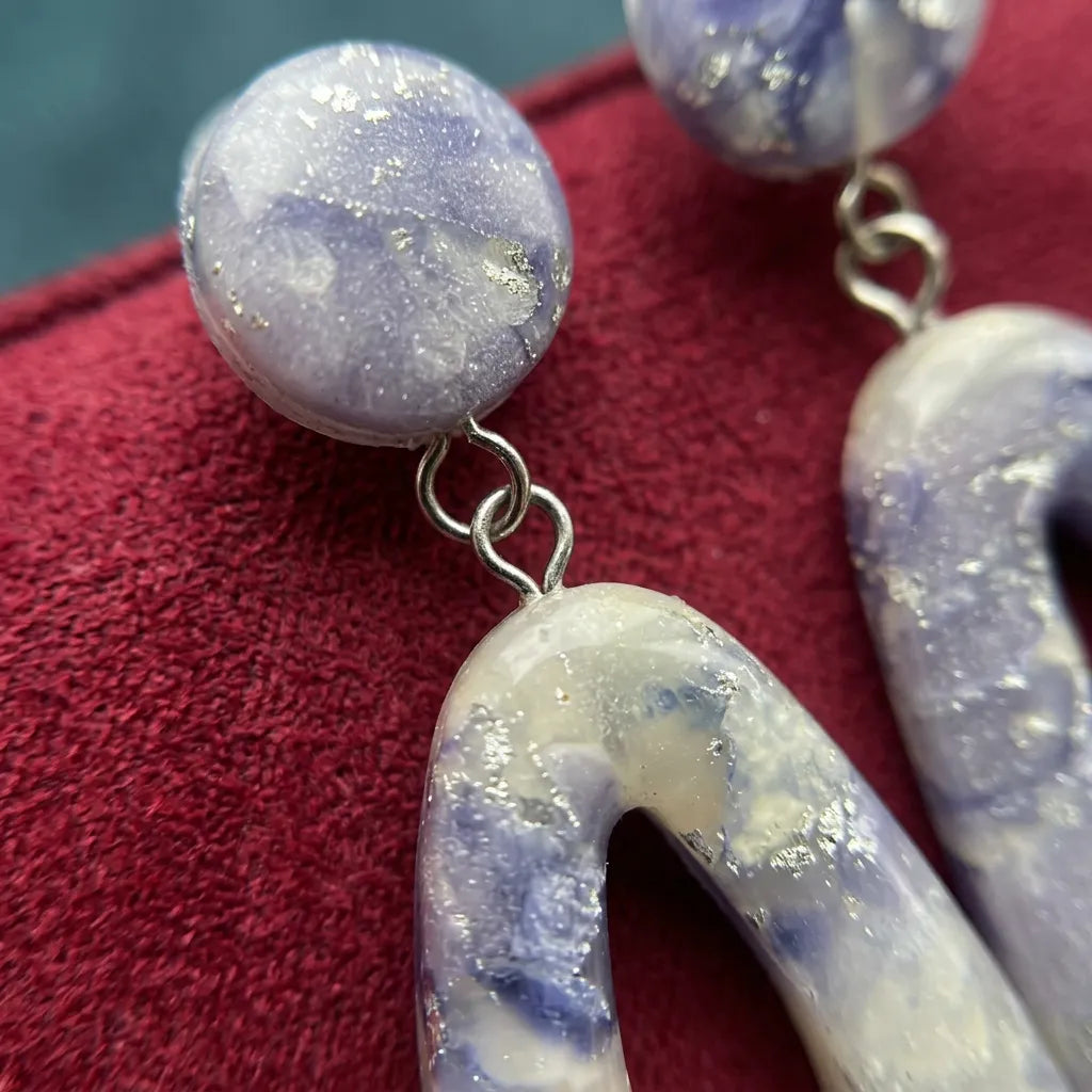"Close-up of a pair of Diddit Amethyst dangling earrings featuring round lavender-blue beads and chunky curved glitter-filled white beads, resting on a red velvet surface."
