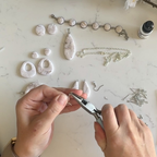 "Hands using silver pliers to assemble a Diddit Mother of Pearl jewellery making experience kit, featuring a white and grey marbled pendant, earrings, and a beaded bracelet laid out on a white marble surface.