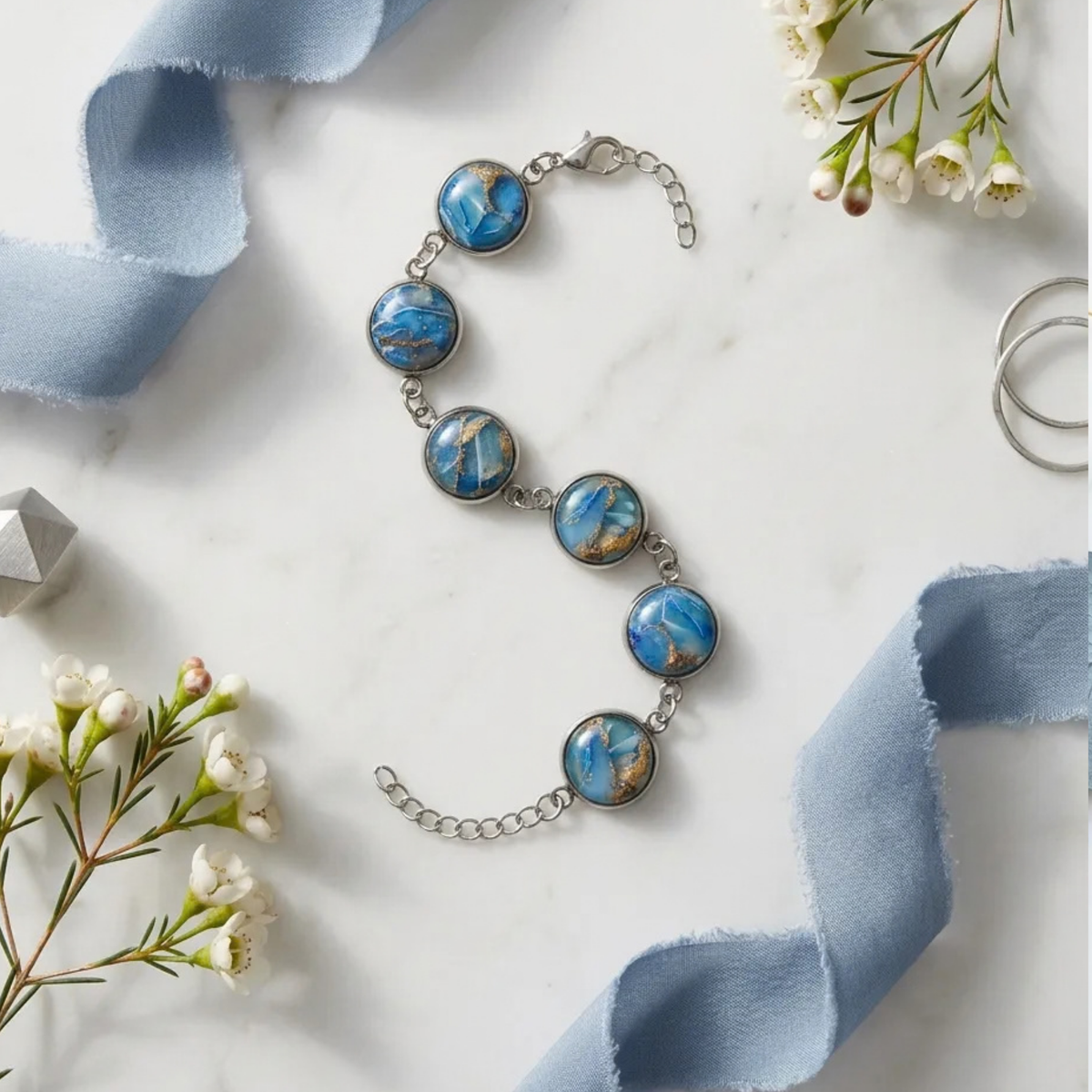 "A Diddit Ocean Effect silver chain bracelet featuring five oval blue and gold marbled stone cabochons, styled on a white marble surface with light blue ribbon and small white flowers."