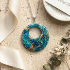 "A Diddit Ocean Effect circular pendant necklace with a marbled blue and turquoise pattern and embedded gold flakes on a silver chain, styled on a textured beige surface with ribbon, a ceramic dish, and small dried flowers."

