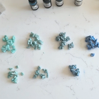 "Six small piles of Diddit Ocean Effect polymer clay pieces in different shades of blue and green arranged on a white marble surface, with three small Diddit alcohol ink bottles visible in the background."