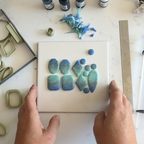 "A pair of hands holding a white tile displaying Diddit Ocean Effect blue and green marbled polymer clay pieces in various shapes, surrounded by olive-green clay cutters, Diddit alcohol ink bottles, and a sanding block on a white marble surface."