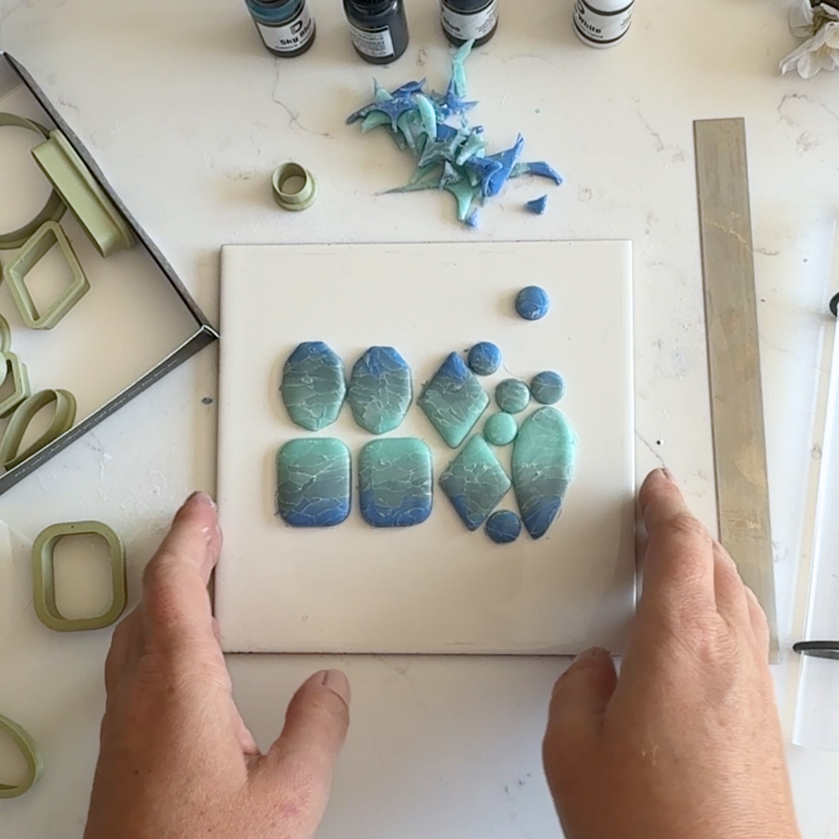 "A pair of hands holding a white tile displaying Diddit Ocean Effect blue and green marbled polymer clay pieces in various shapes, surrounded by olive-green clay cutters, Diddit alcohol ink bottles, and a sanding block on a white marble surface."
