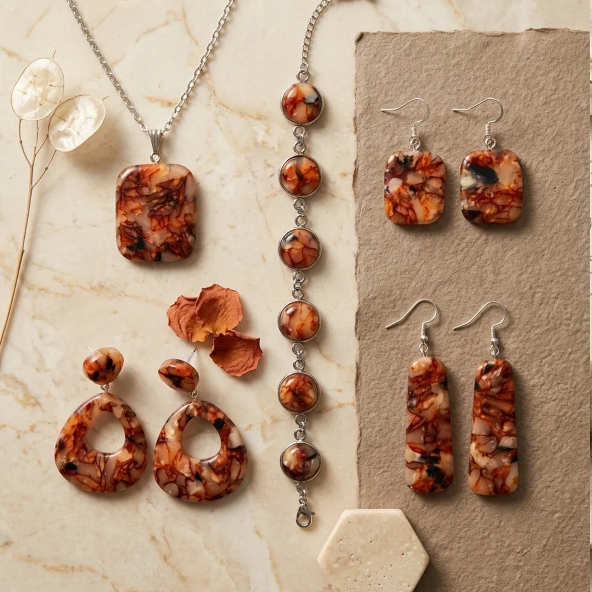 "A Diddit Tortoiseshell jewellery collection flat-lay on a light marble surface, featuring a rectangular pendant necklace, a silver chain bracelet with round cabochons, open teardrop drop earrings, square drop earrings, and long rectangular drop earrings, all crafted from marbled orange, cream, and black tortoiseshell patterned stone with silver-tone findings, styled with dried flowers and petals."