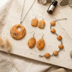 "A Diddit Amber Jewellery Making Experience Kit set displayed on a light stone surface with neutral fabric and dried botanical props, featuring a silver chain necklace with a round orange marbled pendant, matching drop earrings, and a beaded bracelet with orange stone beads and silver links."