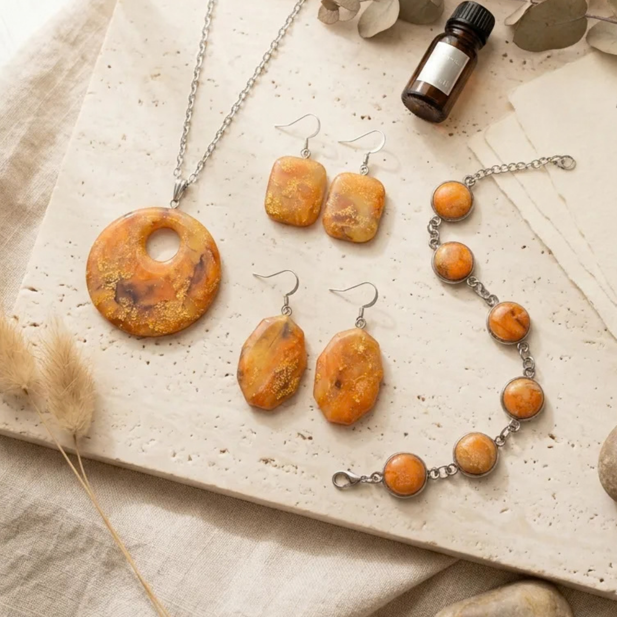 "A Diddit Amber Jewellery Making Experience Kit set displayed on a light stone surface with neutral fabric and dried botanical props, featuring a silver chain necklace with a round orange marbled pendant, matching drop earrings, and a beaded bracelet with orange stone beads and silver links."