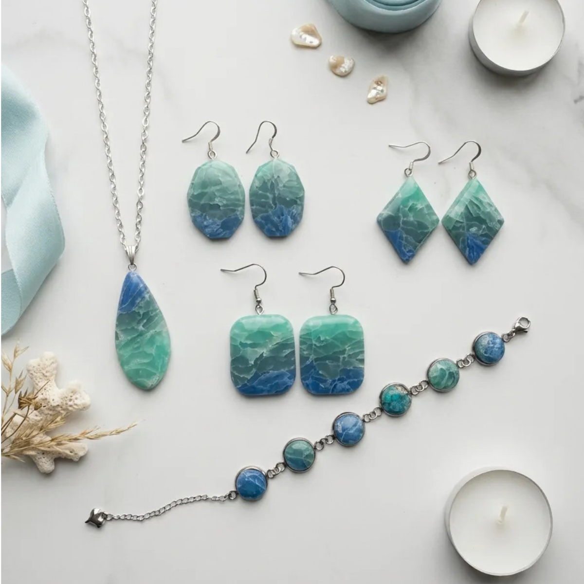 "A Diddit Ocean Effect Jewellery Making Experience Kit set arranged on a light marble surface, featuring a silver teardrop pendant necklace, matching drop earrings, diamond-shaped earrings, square drop earrings, and a beaded bracelet, all with marbled blue and green stones and silver hooks and chain details."
