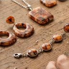 "A Diddit Tortoiseshell jewellery set featuring a pendant necklace, matching earrings, and a bracelet with round and square marbled brown and cream stone pieces set in silver, displayed on a rustic wooden surface."


