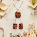 "A Diddit Tortoiseshell jewellery making experience kit set featuring a silver chain necklace and matching drop earrings with rectangular cabochon stones in a marbled orange, brown, and black pattern, styled on a light marble surface with dried flowers and petals."